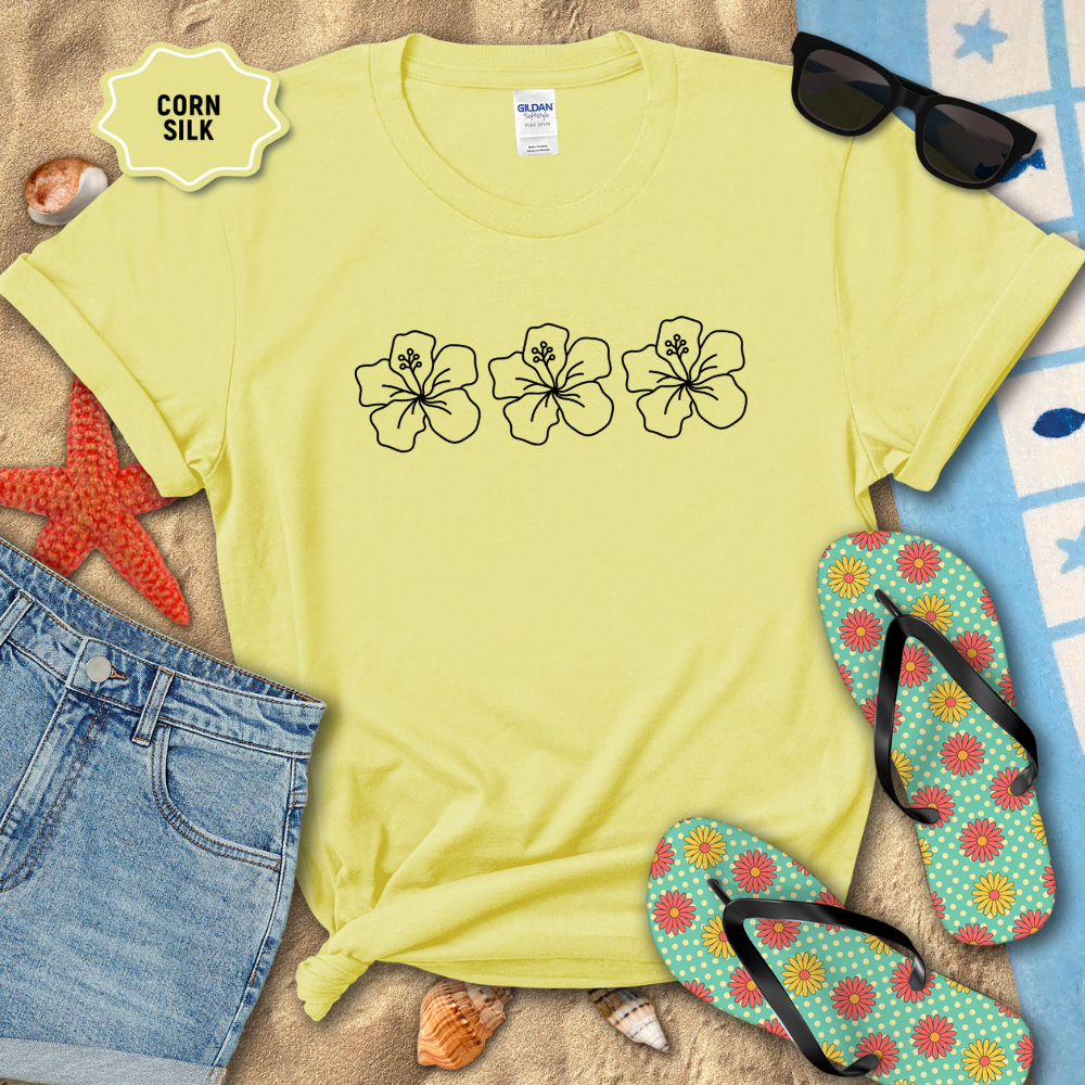 Yellow t-shirt with floral designs on a sandy surface with flip-flops and sunglasses.