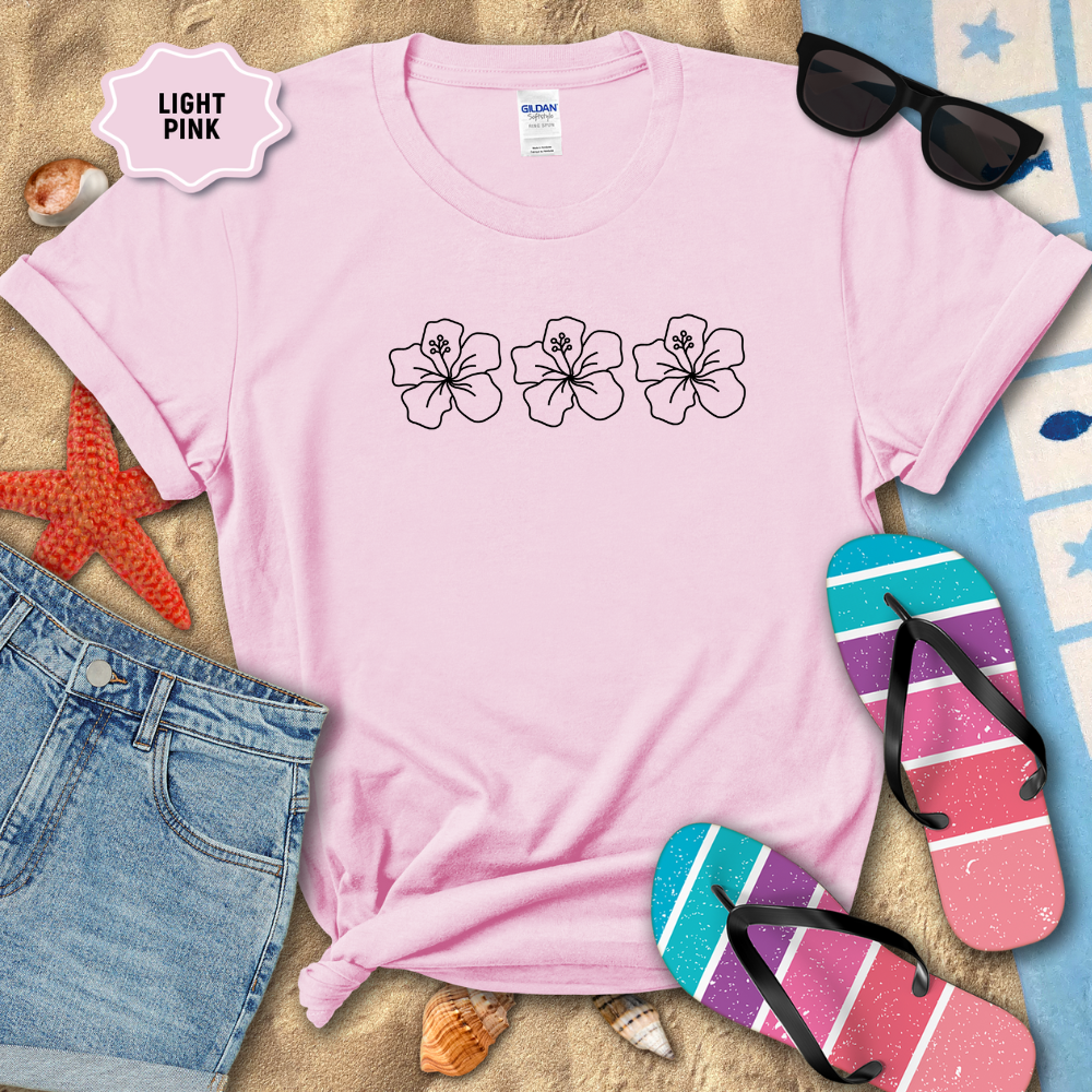 Light pink t-shirt with floral designs on a sandy surface with beach accessories.