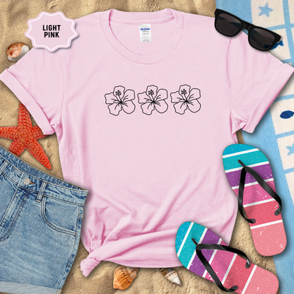 Light pink t-shirt with floral designs on a sandy surface with beach accessories.
