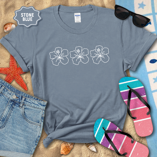 Stone blue t-shirt with floral designs on a beach-themed background with sunglasses, flip-flops, and denim shorts.