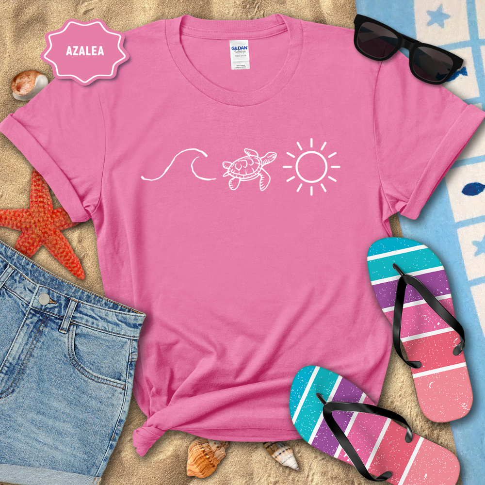 Pink t-shirt with beach-themed design on a sandy surface with flip-flops and sunglasses.