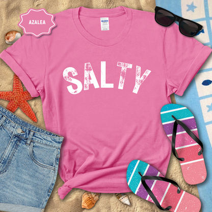 Pink t-shirt with 'SALTY' text on a beach-themed background with sunglasses, flip-flops, and denim shorts.