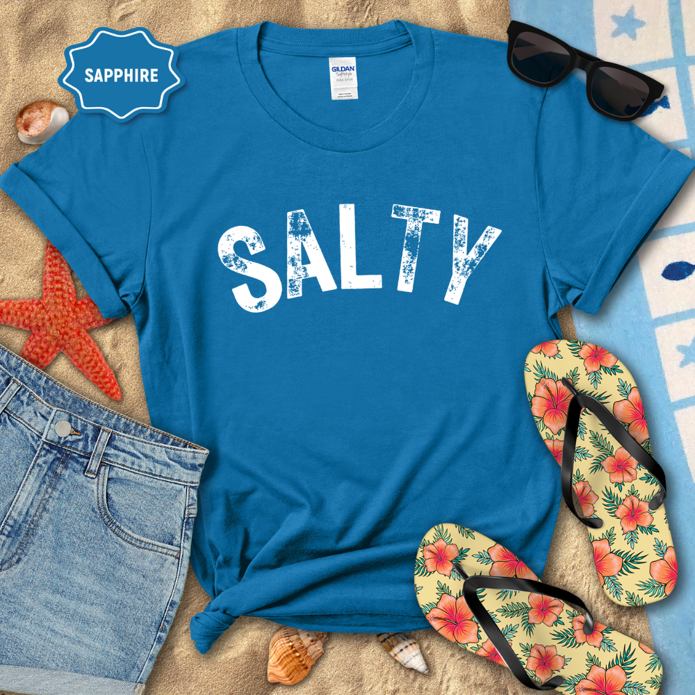 Blue t-shirt with 'SALTY' text, sunglasses, flip-flops, and beach items on a sandy background.