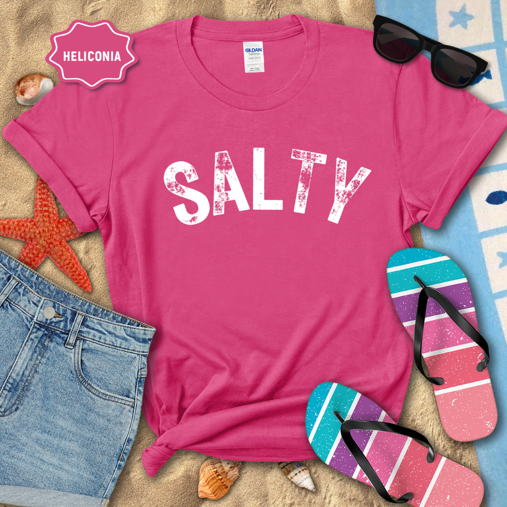 Pink t-shirt with 'SALTY' text on a sandy surface with beach accessories.