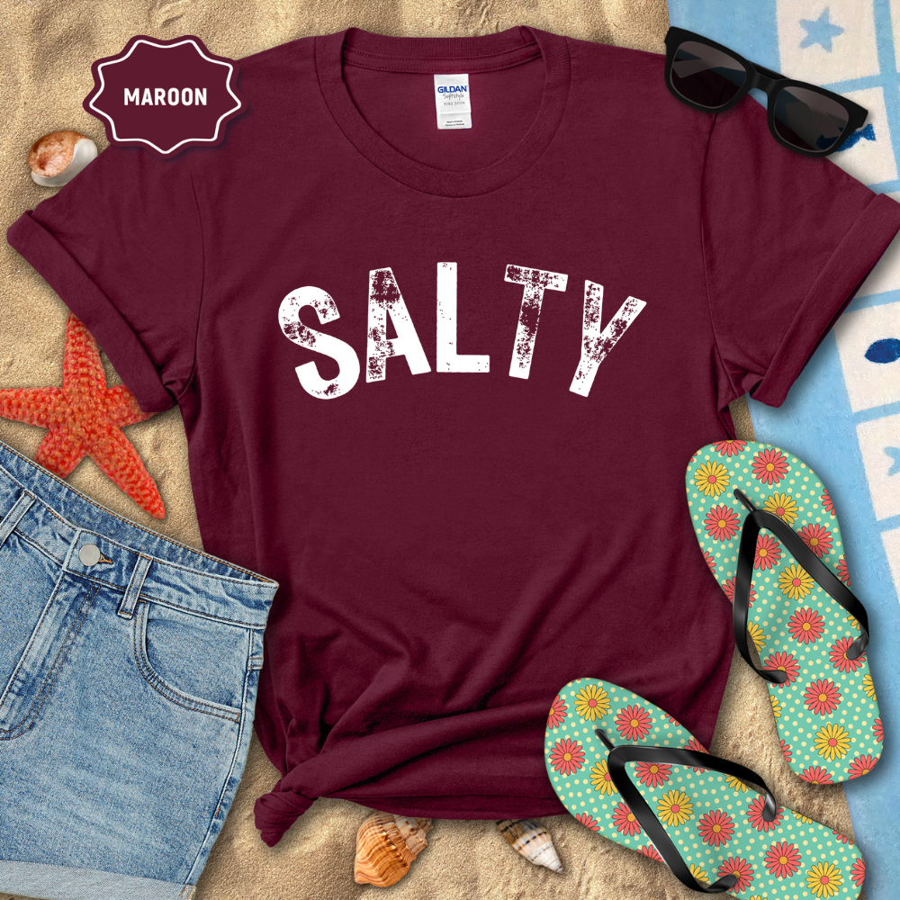 Maroon t-shirt with 'SALTY' text on a sandy surface with beach accessories.