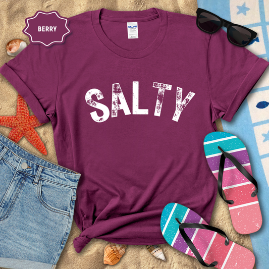 Purple t-shirt with 'SALTY' text, sunglasses, denim shorts, and flip-flops on a beach-themed background.