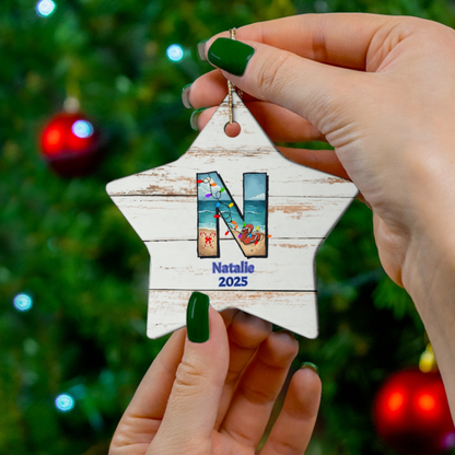 Hand holding a star-shaped Christmas ornament with 'Natalie 2025' against a blurred Christmas tree background.