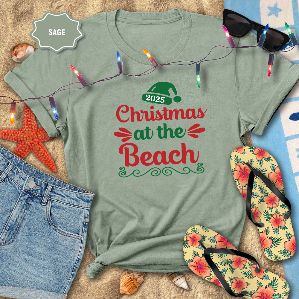 Sage green t-shirt with 'Christmas at the Beach 2025' design, surrounded by beach-themed items.