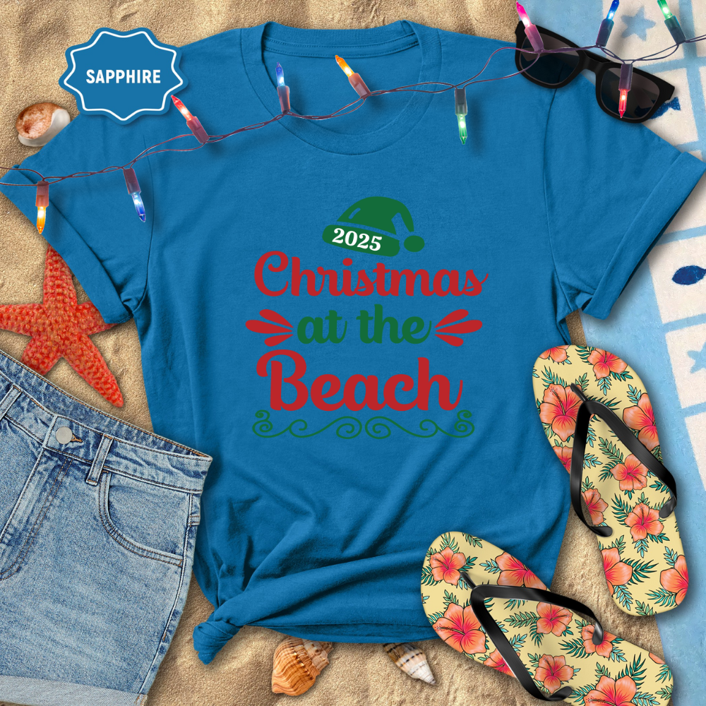 Blue t-shirt with '2025 Christmas at the Beach' text on a sandy background with beach accessories.