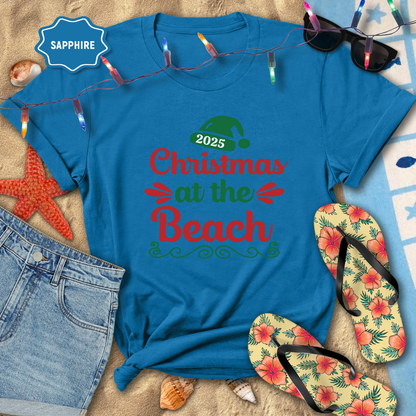 Blue t-shirt with '2025 Christmas at the Beach' text on a sandy background with beach accessories.