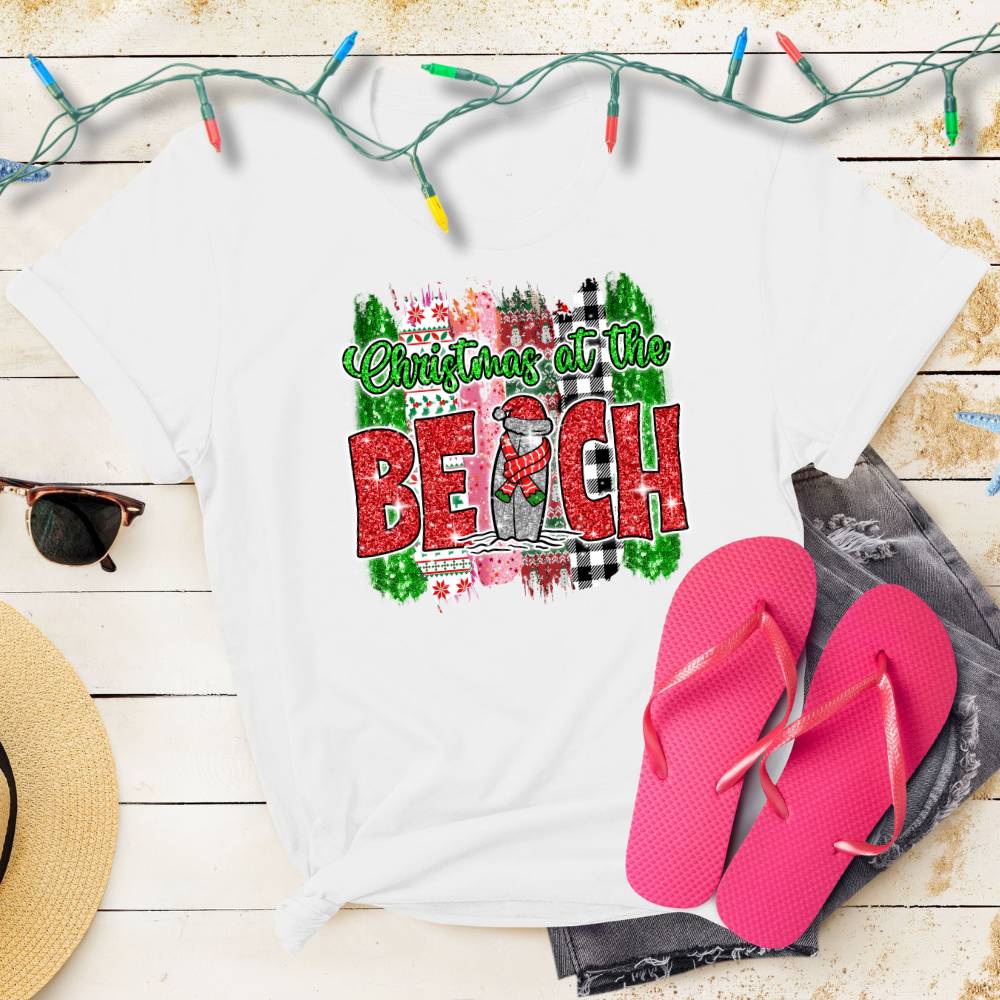 White t-shirt with 'Christmas at the Beach' design, pink flip-flops, and Christmas lights on a wooden surface.
