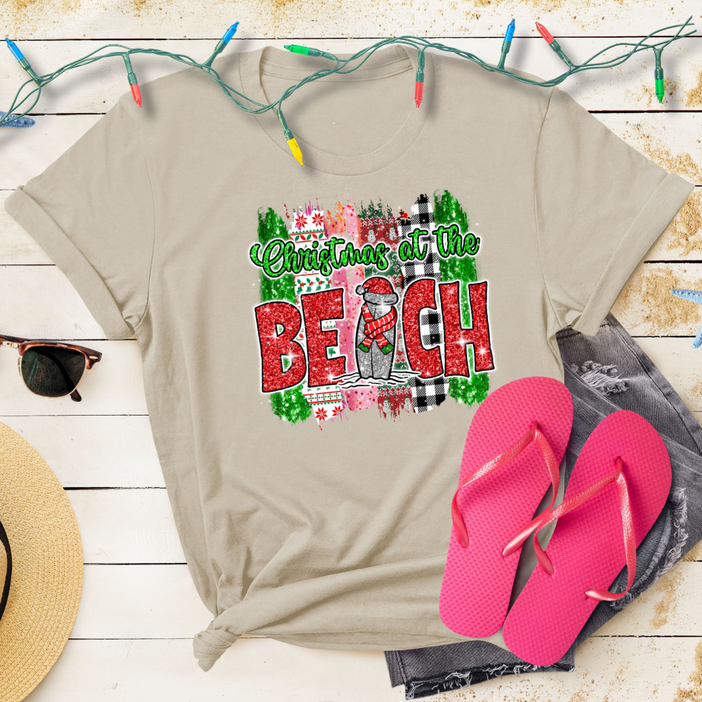 Beige t-shirt with 'Christmas at the Beach' design, pink flip-flops, and Christmas lights on a wooden surface.