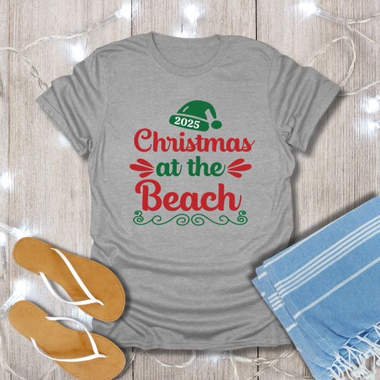 Gray t-shirt with 'Christmas at the Beach 2025' text, flip-flops, and towel on a wooden surface.