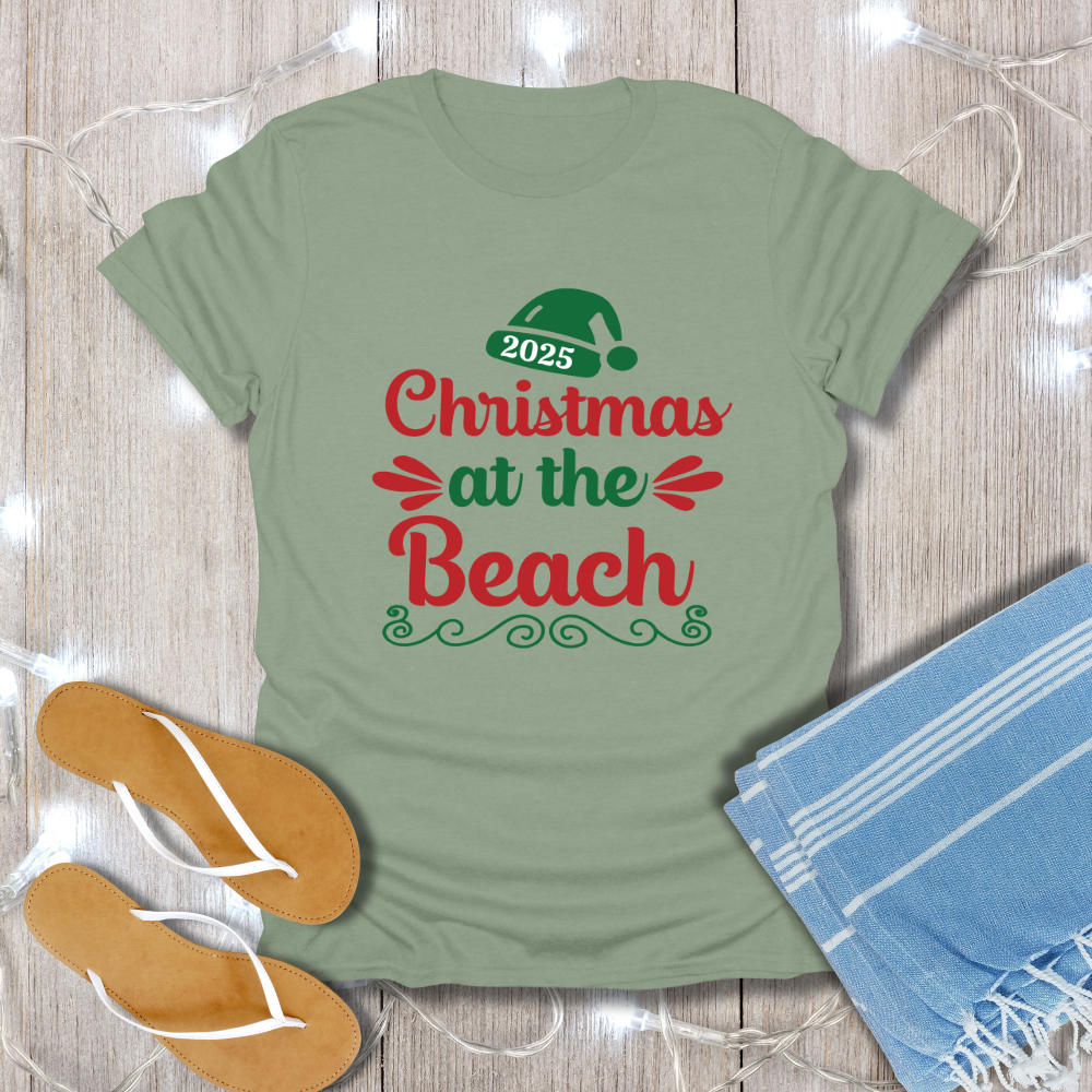 Green t-shirt with 'Christmas at the Beach 2025' text on a wooden surface with flip-flops and a towel.