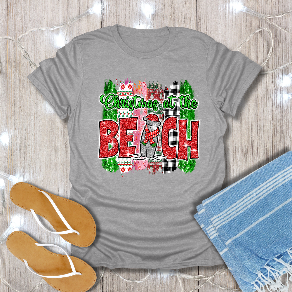 Gray t-shirt with 'Christmas at the Beach' design on a wooden surface with flip-flops and a towel.