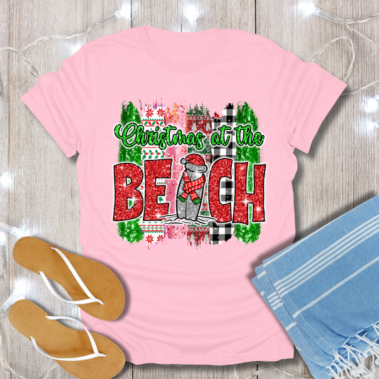 Pink t-shirt with 'Christmas at the Beach' design on a wooden surface with flip-flops and a towel.