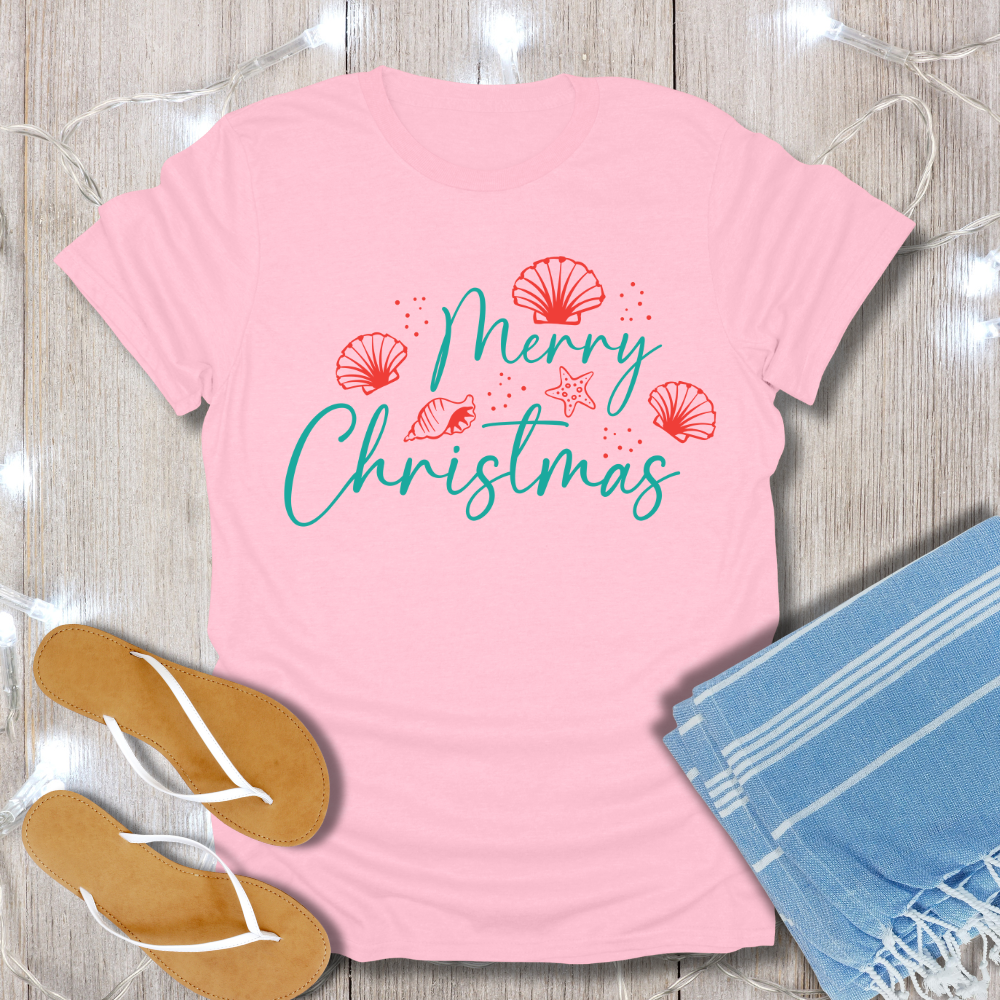 Pink t-shirt with 'Merry Christmas' text and decorative elements on a wooden surface with flip-flops and a towel.