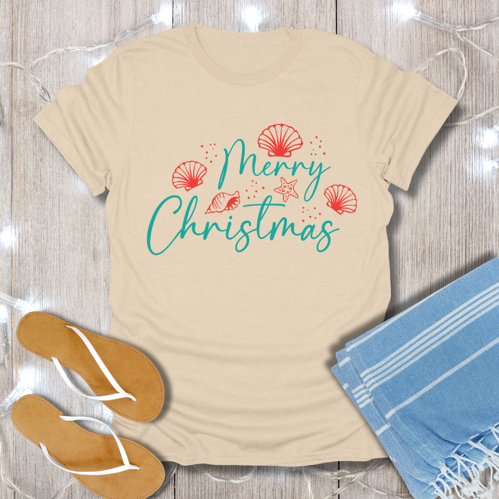 Beige t-shirt with 'Merry Christmas' text and decorative elements on a wooden surface with flip-flops and a towel.