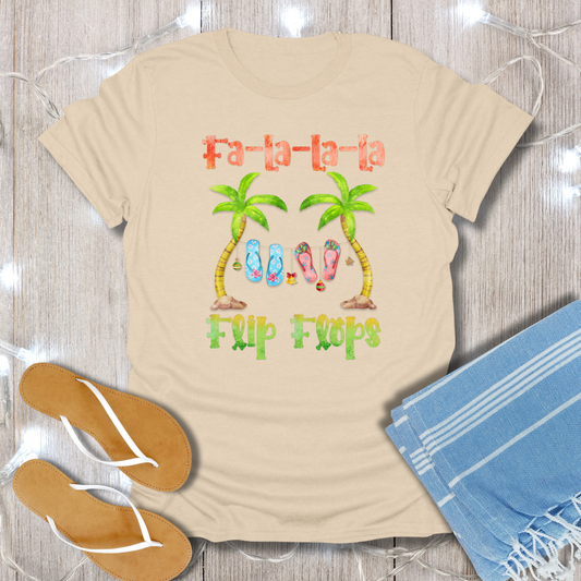 Beige t-shirt with 'Fa-la-la-la Flip Flops' design on a wooden surface with flip-flops and a towel.