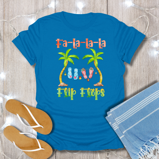 Blue t-shirt with 'Fa-la-la-la Flip Flops' design on a wooden surface with flip flops and a towel.