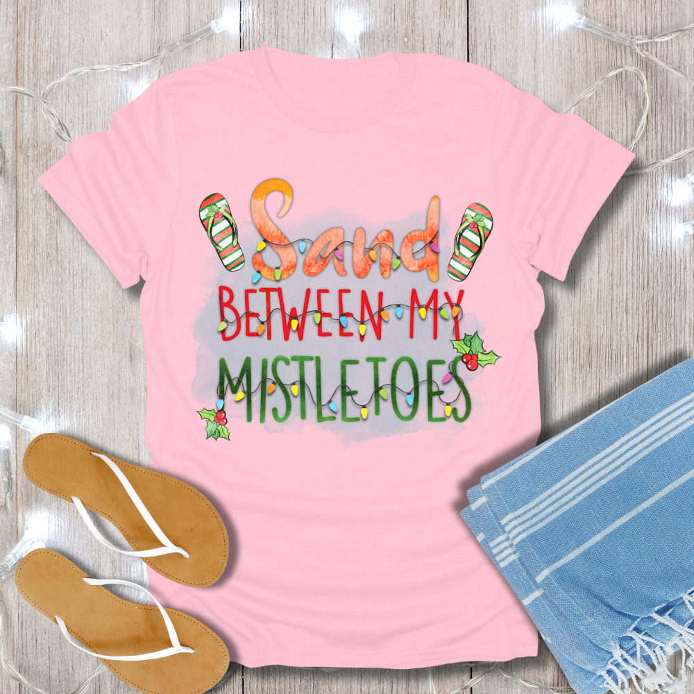 Pink t-shirt with 'Sand Between My Mistletoes' text, flip-flops, and towel on a wooden surface.