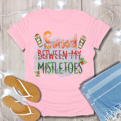 Pink t-shirt with 'Sand Between My Mistletoes' text, flip-flops, and towel on a wooden surface.