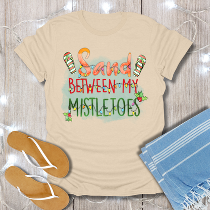 Beige t-shirt with 'Sand Between My Mistletoes' text, flip-flops, and towel on a wooden surface.
