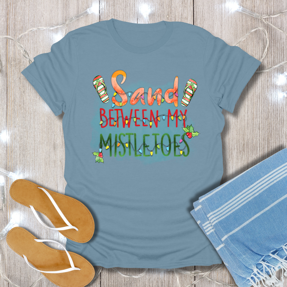 Blue t-shirt with 'Sand Between My Mistletoes' text on a wooden surface with flip-flops and a towel.