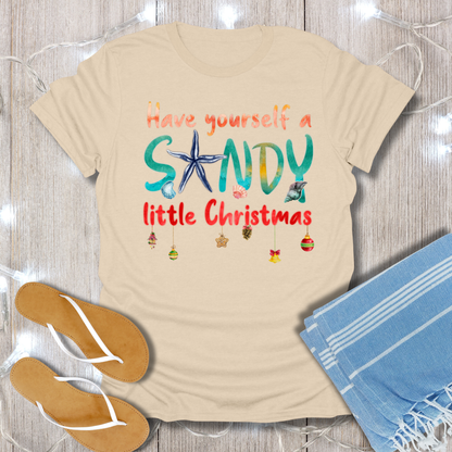 Beige t-shirt with 'Have yourself a SANDY little Christmas' text and beach-themed graphics on a wooden surface.