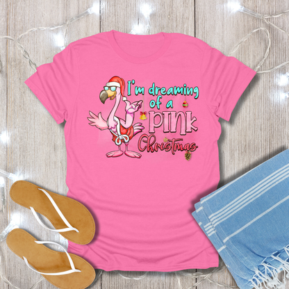 Pink t-shirt with flamingo graphic and text 'I'm dreaming of a pink Christmas' on a wooden surface with flip-flops and a towel.
