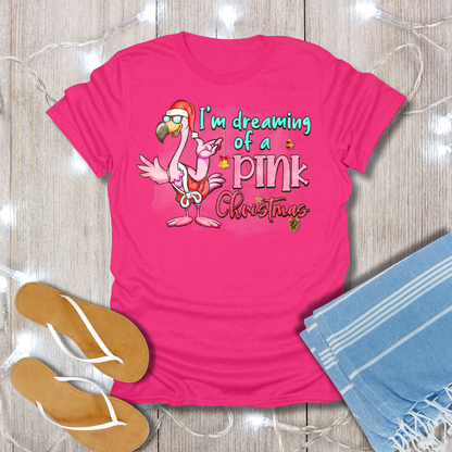 Pink t-shirt with flamingo graphic and text 'I'm dreaming of a pink Christmas' on a wooden surface with flip-flops and a towel.