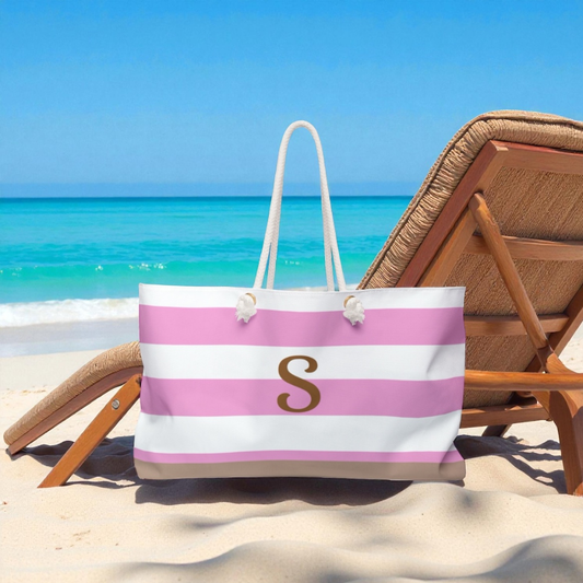 Beach bag with pink and white stripes and a gold 'S' on a sandy beach with a chair and ocean view.