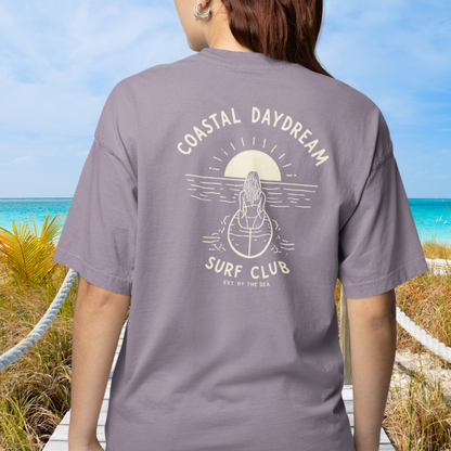 Person wearing a gray t-shirt with 'Coastal Daydream Surf Club' design on a beach.