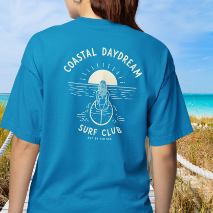 Person wearing a blue t-shirt with 'Coastal Daydream Surf Club' design on a beach.