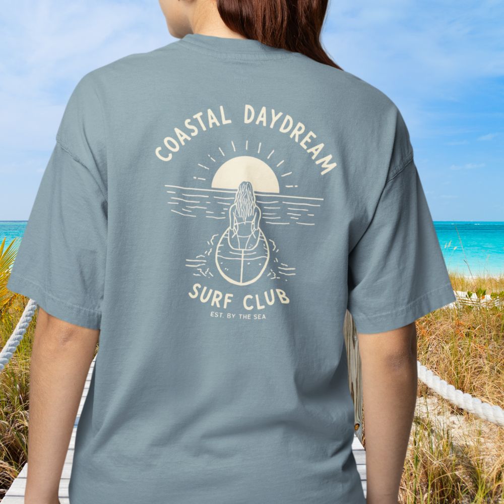 Person wearing a gray t-shirt with 'Coastal Daydream Surf Club' design on a beach.