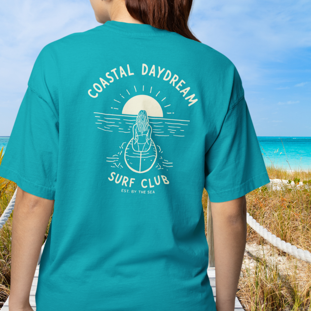 Person wearing a teal t-shirt with 'Coastal Daydream Surf Club' design on a beach.