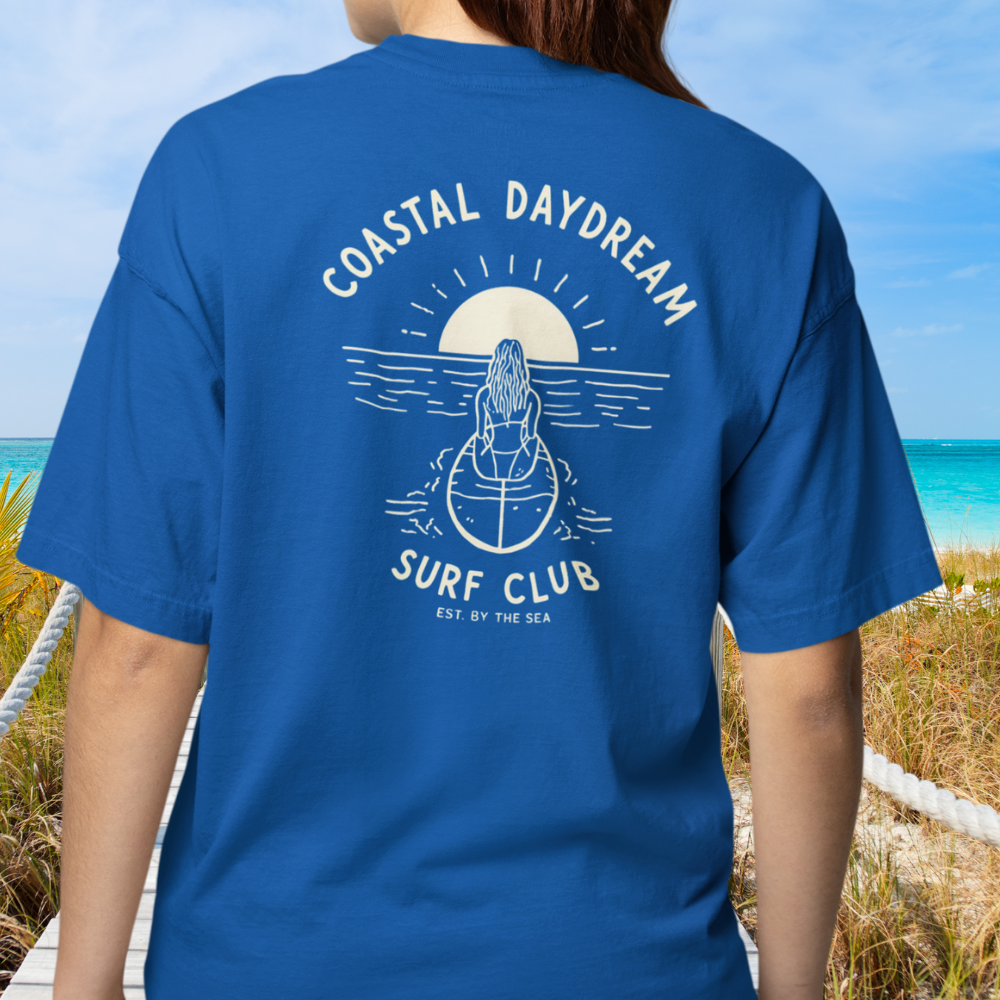 Person wearing a blue t-shirt with 'Coastal Daydream Surf Club' design on a beach.
