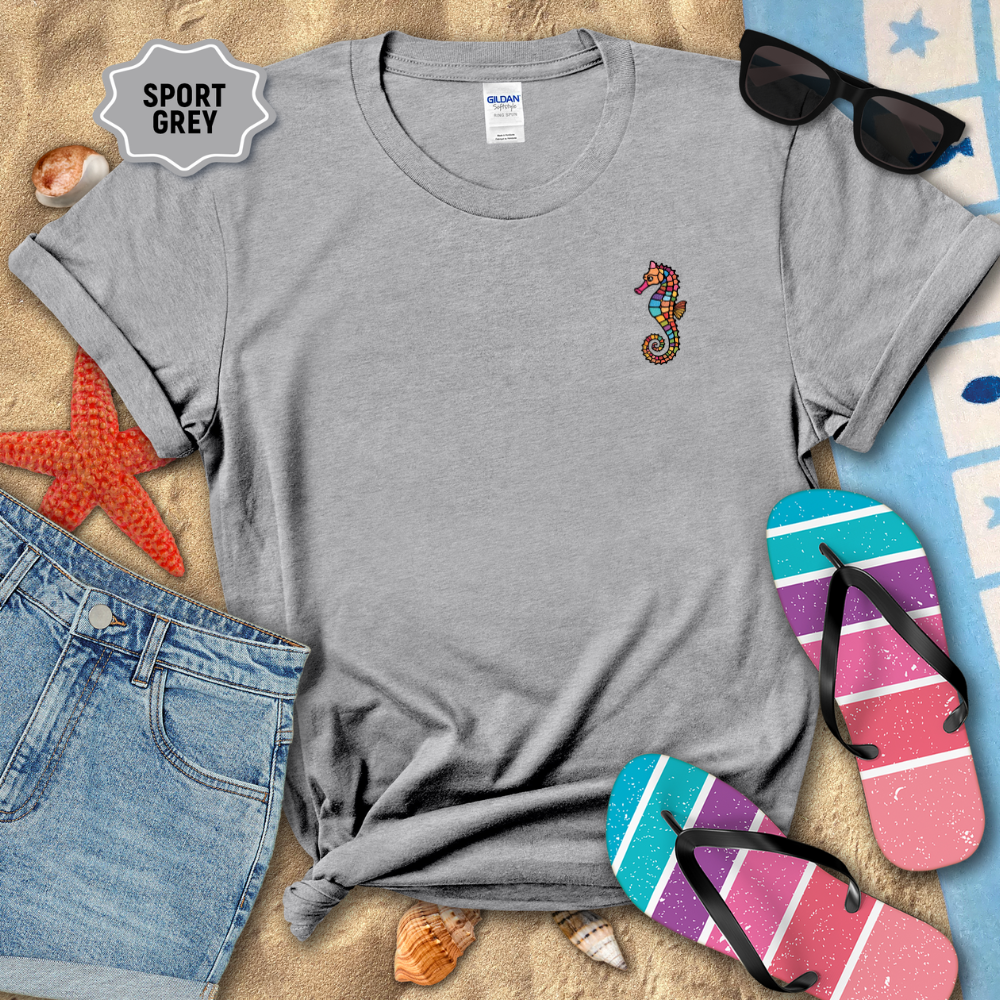 Gray t-shirt with a colorful seahorse design on a sandy surface with beach accessories.