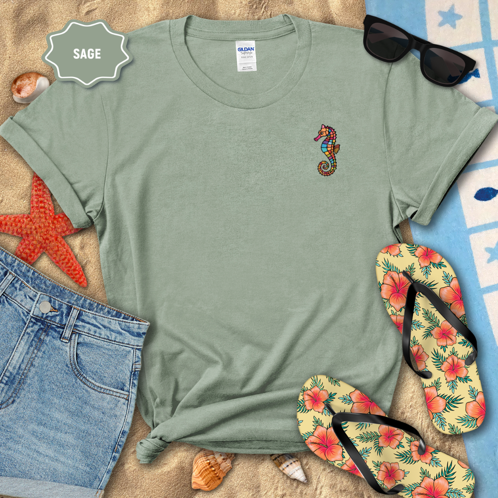 Sage green t-shirt with a seahorse design, sunglasses, flip-flops, and beach items on sand.