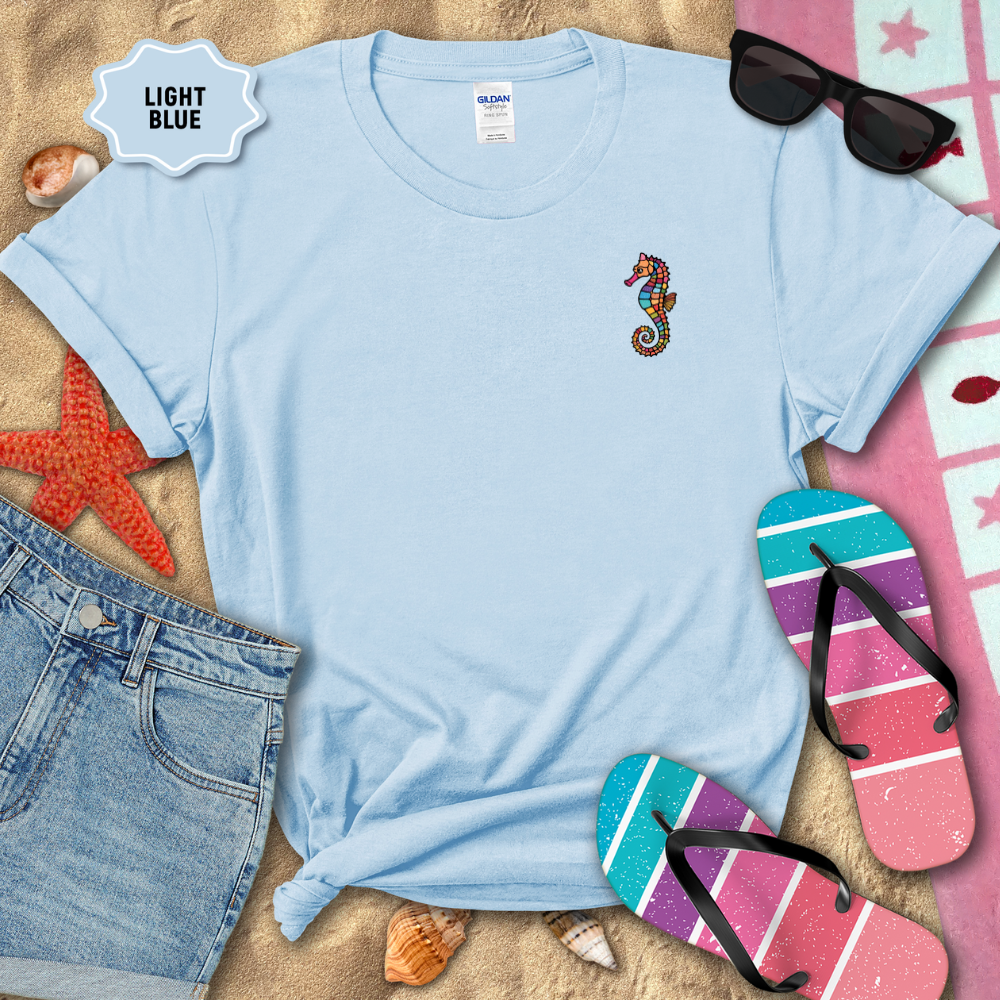 Light blue t-shirt with a seahorse design on a sandy background with beach accessories.