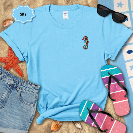 Blue t-shirt with a seahorse design on a sandy surface with beach accessories.