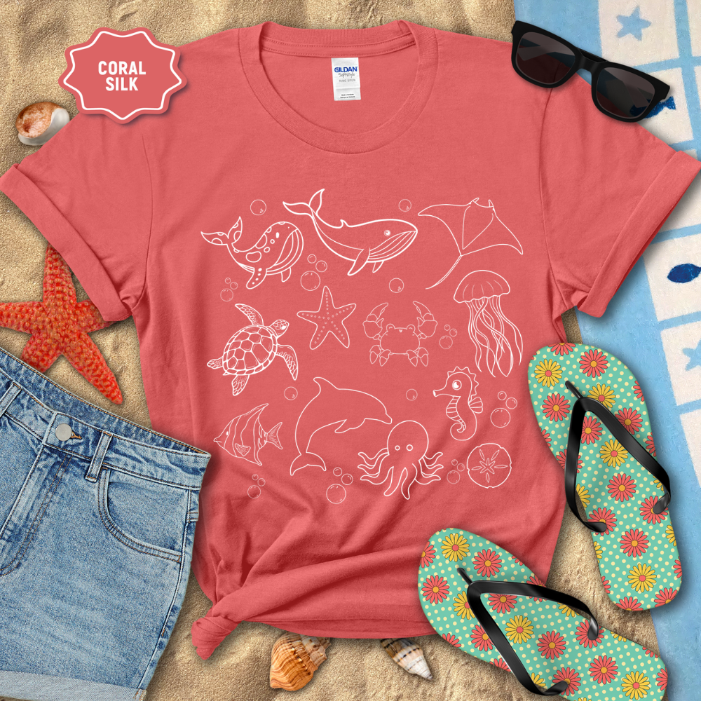 Coral t-shirt with white sea creature designs on a sandy surface with sunglasses and flip-flops.