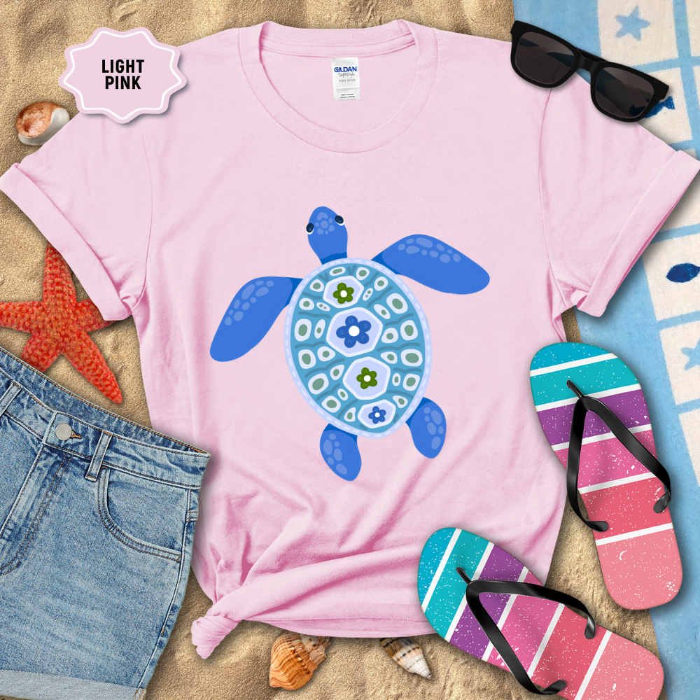 Light pink t-shirt with blue turtle design on a sandy surface with beach accessories.