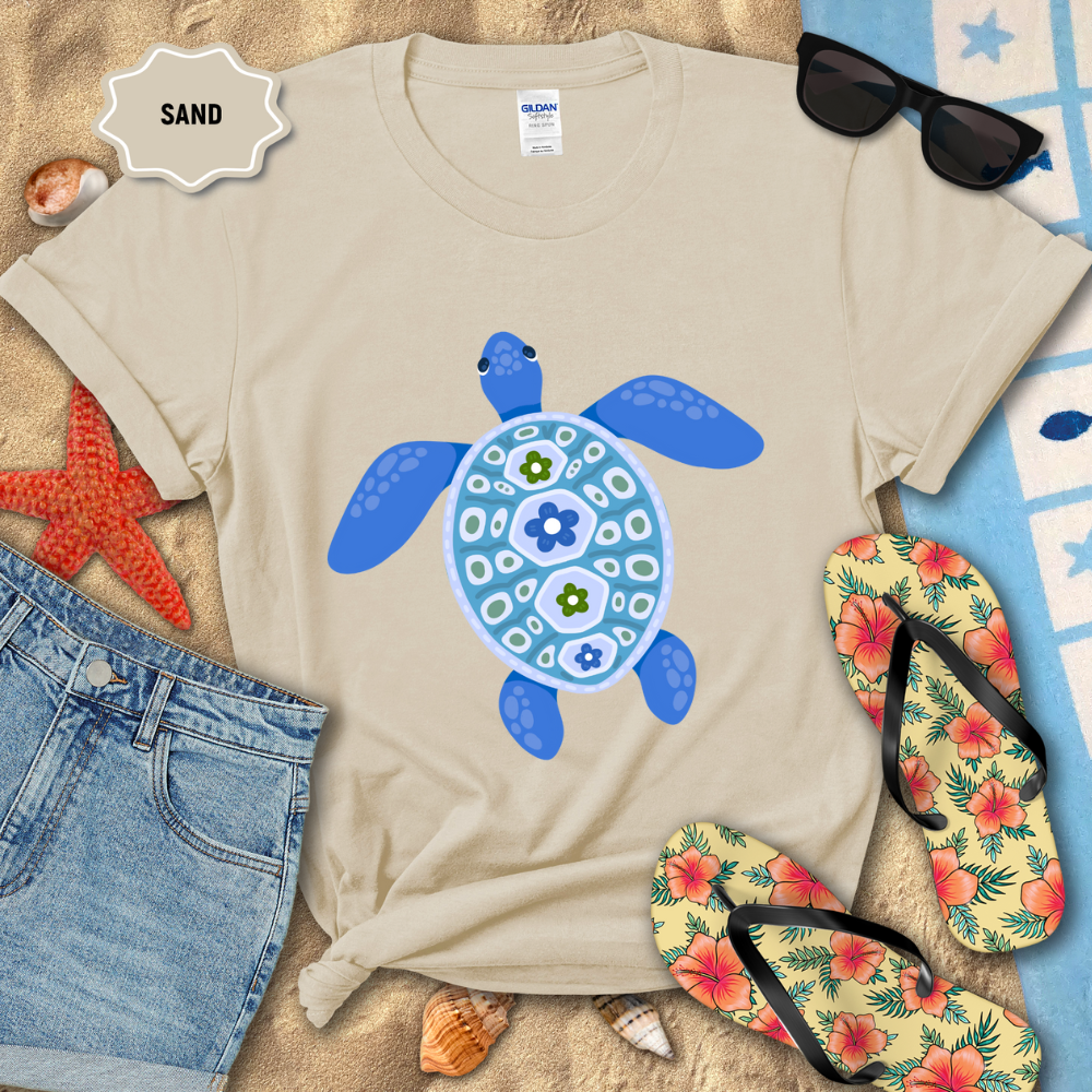 Beige t-shirt with blue turtle design on sand with sunglasses, flip-flops, and starfish.