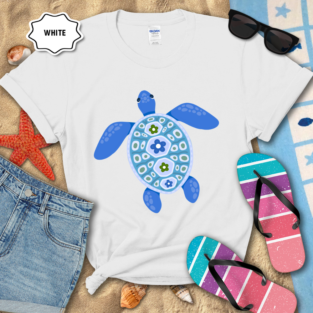 White t-shirt with a blue turtle design on a sandy surface with flip-flops and sunglasses.