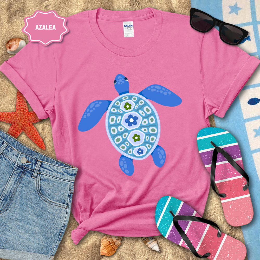 Pink t-shirt with a blue turtle design on a sandy surface with beach accessories.