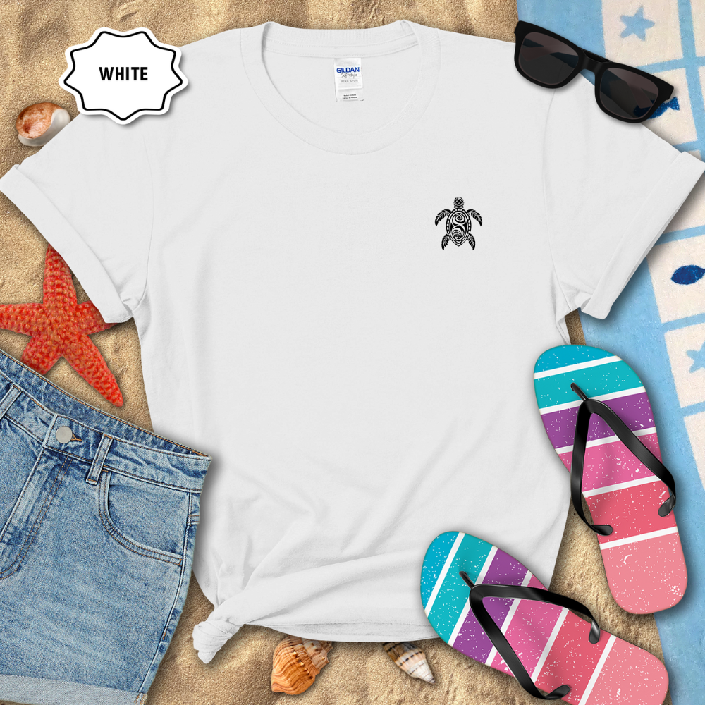 White t-shirt with a turtle design on a beach setting with sunglasses, flip-flops, and denim shorts.