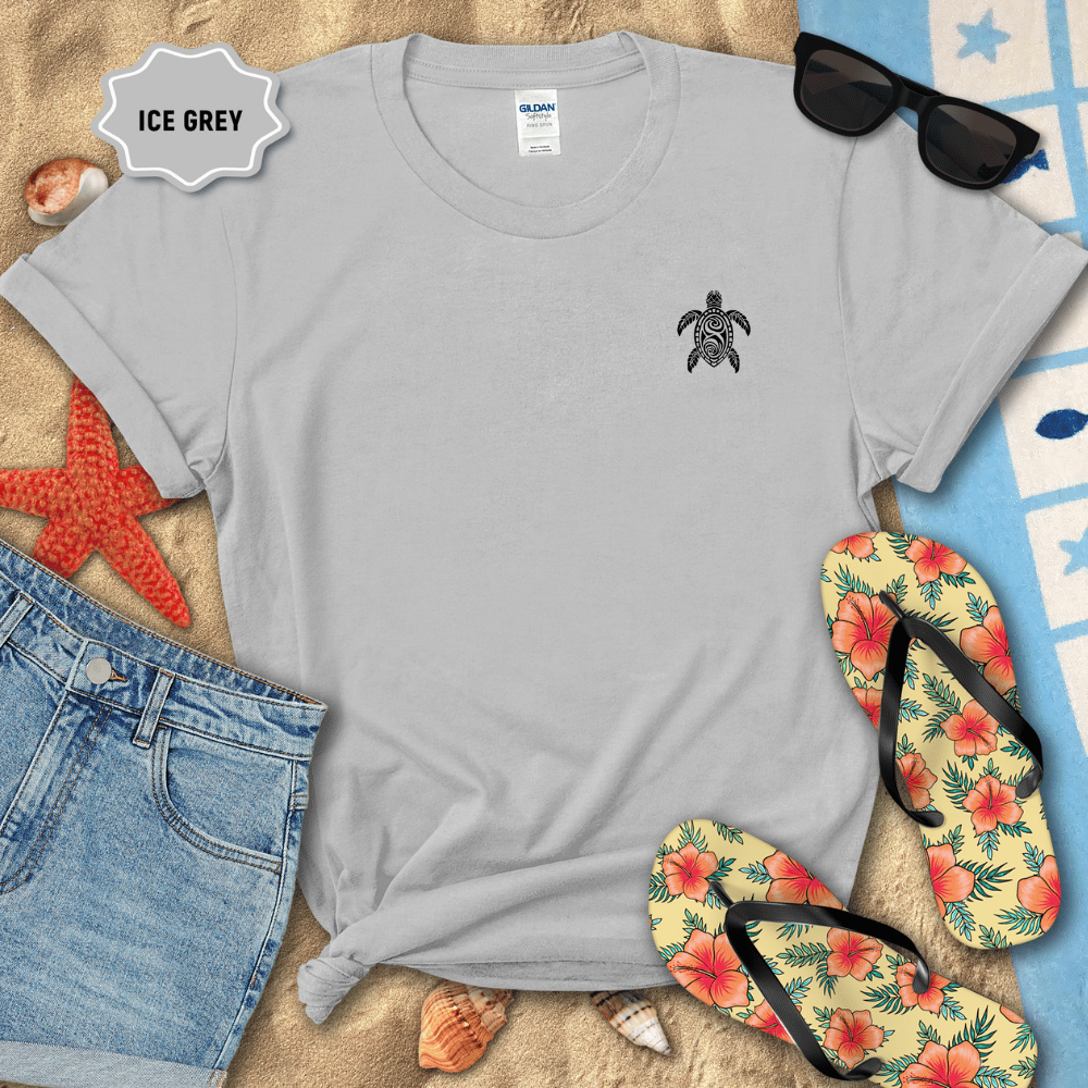 Gray t-shirt with a turtle design on a sandy surface with beach accessories.