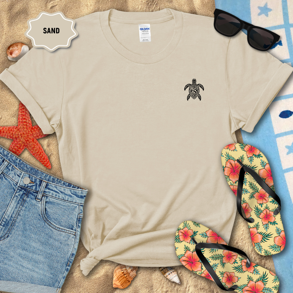Beige t-shirt with a turtle design on a sandy background with sunglasses, flip-flops, and beach accessories.