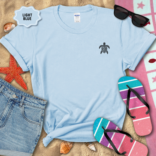 Light blue t-shirt with a turtle design on a sandy background with beach accessories.